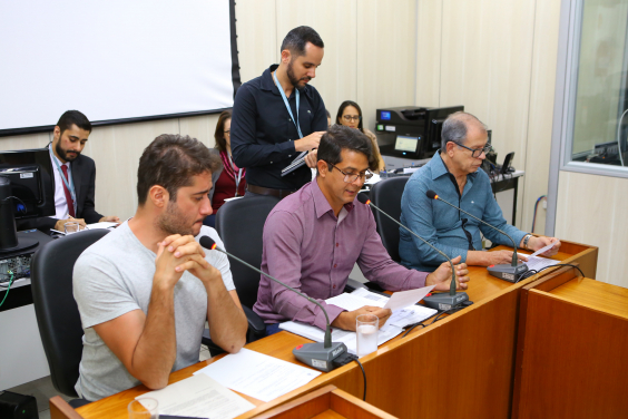 Três integrantes da comissão estão sentados à Mesa do Plenário Camil Caram; um servidor de pé e três assentados atrás deles assessoram e registram as ocorrências da reunião.