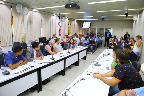 Vista aberta do plenário. Parlamentares e convidados compõem mesa de reunião. Ao fundo, pessoas acompanham a reunião.