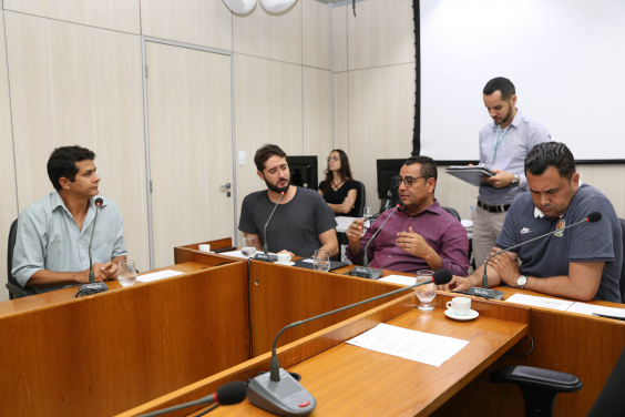 Integrantes da Comissão estão sentados à Mesa, diante dos respectivos microfones apreciando a pauta da reunião. Atrás deles, um coordenador do processo legislativo está de pé, assessorando a reunião