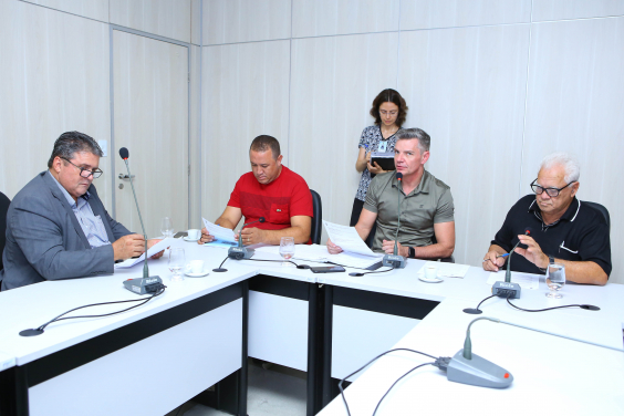 Integrantes da Comissão estão sentados à Mesa, diante de microfones, apreciando a pauta da reunião; atrás deles,