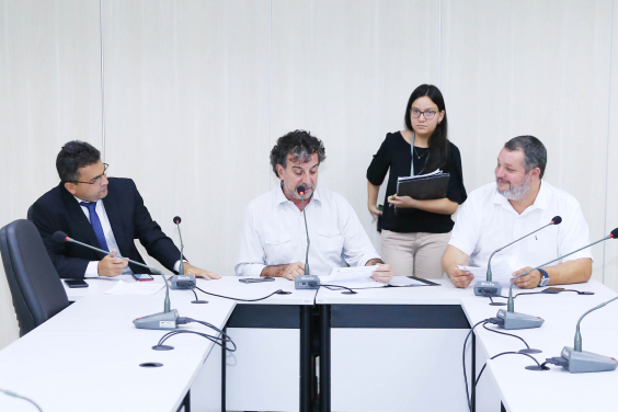 Integrantes da Comissão estão sentados à Mesa apreciando a pauta da reunião. Atrás deles, de pé, uma servidora assessora o presidente.