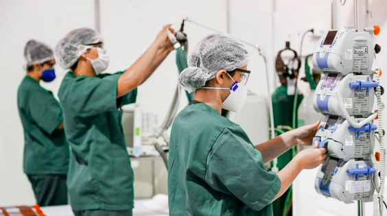 a imagem mostra três enfermeiros de pé, com jalecos verdes, operando equipamentos em UTI