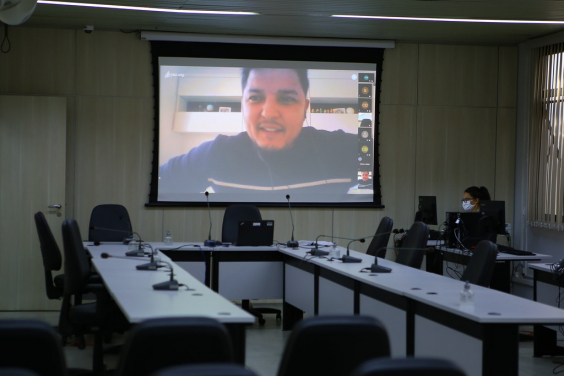 A imagem mostra a mesa do plenário vazia, com a equipe de assessoria ao lado,  e um telão com as imagens dos vereadores em participação remota