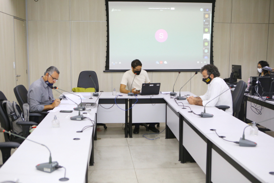 Três vereadores sentados à mesa e telão ao fundo em videoconferência com convidados