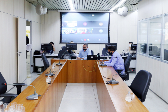 Parlamentares compõem mesa de reunião. Telão ao fundo em videoconferência com os demais membros e convidados da CPI