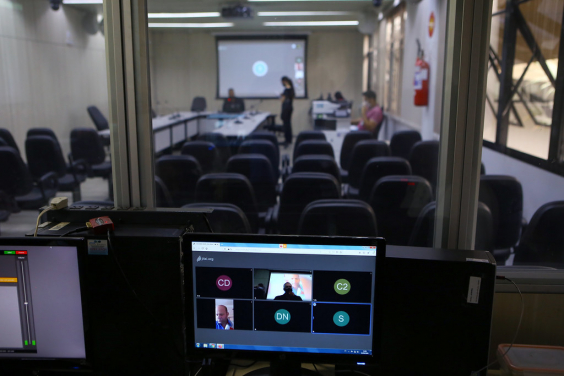 Tela de computador exibindo videoconferencia. Ao fundo, cadeiras vazias no pplnaéio e telão exibindo videoconferência
