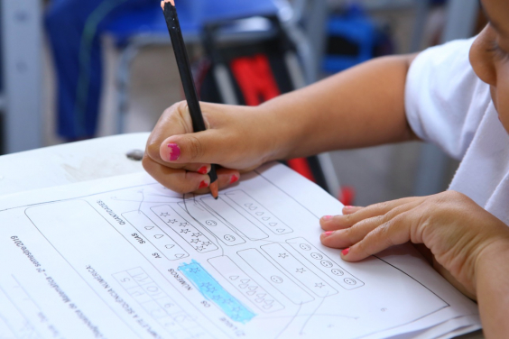 Mãos de criança escrevendo em papel sobre a mesa. Papel exibe atividade escolar