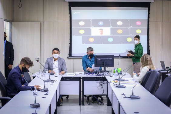 sala de reunião com quatro vereadores sentados distantes um do outro e ao fundo tela de tv com participantes remotos