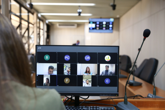 Monitor de computador exibe vereadores em videoconferência. Ao fundo, cadeiras vazias do Plenário Camil Caram