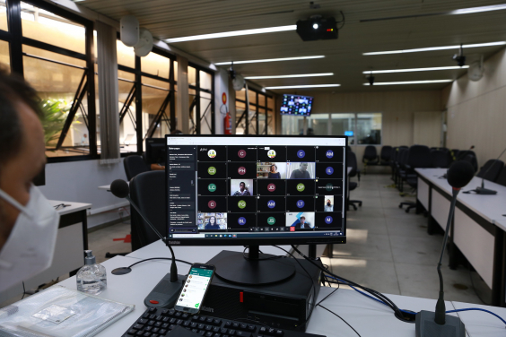 Monitor de computador exibe vereadores em videoconferência. Ao fundo, cadeiras vazias do Plenário Helvécio Arantes