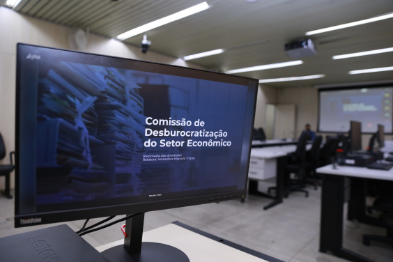 tela de computador em primeiro plano mostra nome da Comissão; a fundo, estrutura do Plenário Camil Caram 