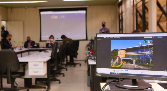 Reunião de Plenário realizada no plenário Helvécio Arantes. Monitor de computador exibe foto da fachada da Câmara. Ao fundo, vereadores compõem mesa de reunião. Telão exibe demais parlamentares em videoconferência