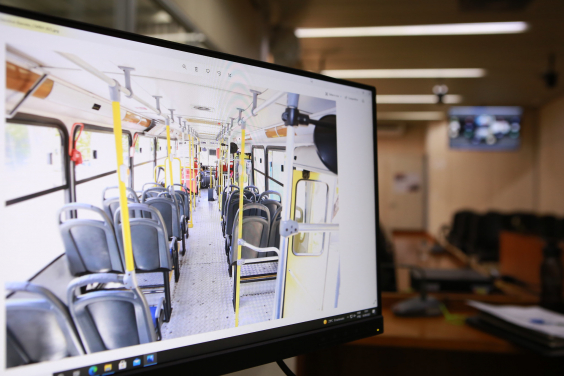 tela de computador, em primeiro plano, mostra interior de um ônibus; ao fundo, mobília do Plenário Camil Caram 