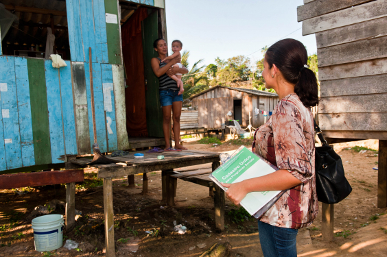 Profissional de assistência social visita área carente da cidade com caderno de anotações do Cadastro único, durante o dia.