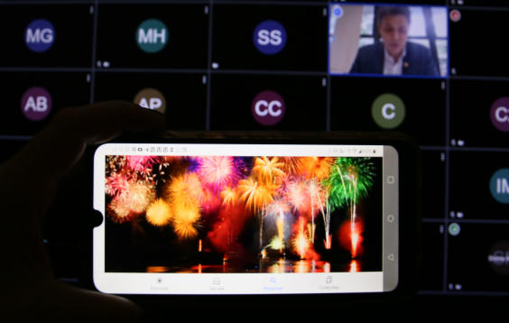 Imagem das luzes dos fogos de artifícios na tela do celular. Ao fundo a tela do comutador com a imagem dos participantes da audiência
