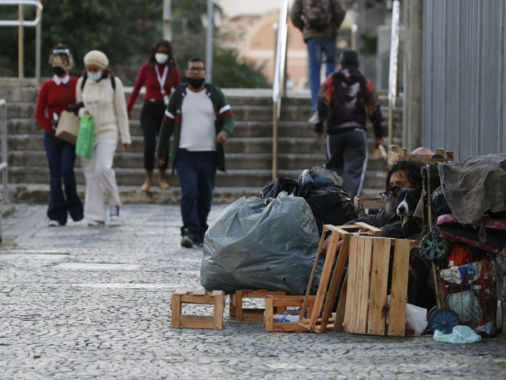 Morador de rua cercado de pertences, sentado em rua de cidade encostado em muro, com seis cidadãos passando ao fundo, próximo a uma escada de seis degraus, durante o dia.