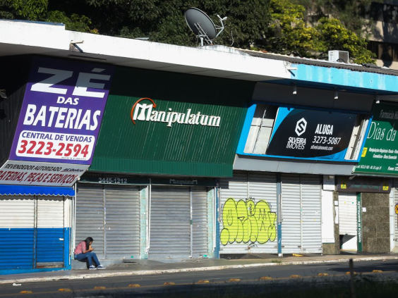 Mulher sentada próxima a quatro lojas com portas fechadas, durante o dia.