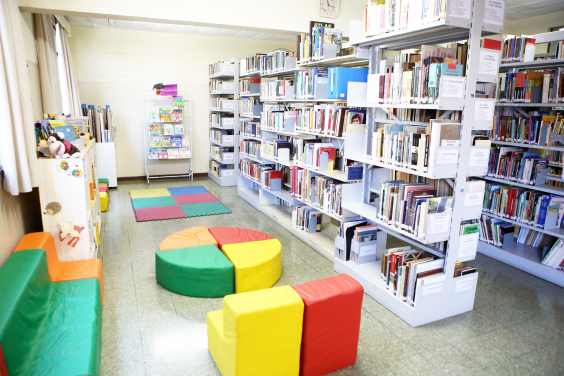 Biblioteca infantil, com bancos e tapetes coloridos. Ao centro, mesas formada por quatro triângulos coloridos