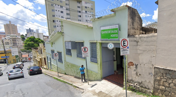 Fachada de Centro de Saúde Horto, com cidadã caminhando em frente, na calçada.  a rua é inclinada, e mais de cinco carros aparecem no final da imagem diurna, em direção contrária à do centro de saúde. 