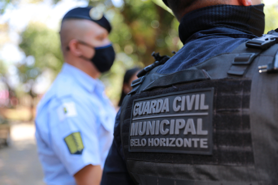 Dois guardas municipais, um de lado e outro de costas, de colete, durante o dia. 