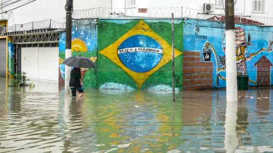 Uma pessoa com guarda-chuva na mão atravessa a rua alagada . Ao fundo, um muro pintado com a bandeira do Brasil onde se lê Ordem e Progresso