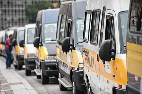 Mais de oito veículos de transporte escolar enfileirados, durante o dia. Ao fundo, um homemn conversa fora do veículo, voltado para a janela de um deles. 