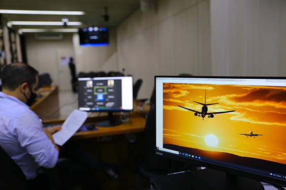 Tela mostra avião sobrevoando a cidade com um céu alaranjado, indicando o pôr-do-sol. Um homem caucasiano , vestido com uma camisa azul de mangas compridas, está sentado ao lado segurando algumas folhas de papel , 