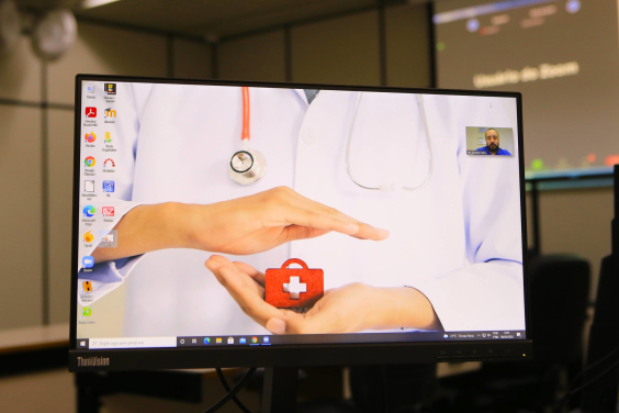 Pessoa vestida com jaleco branco e na palma das mãos, posicionadas à frente do corpo, uma pequena maleta vermelha com uma cruz branca -símbolo do SUS