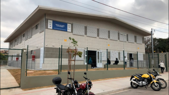 Fachada do Centro de Sa[ude Mantiqueira, uma casa de dois andares, duas portas e seis janelas frontais, seis pessoas à frente, grade e quatro motos na rua à frente, durante o dia.