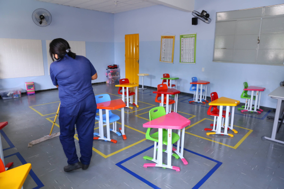 Mulher de conjunto de calça e blusa azul e máscara, de costas, limpa sala de Unidade de Educação Infantil com rodo. 