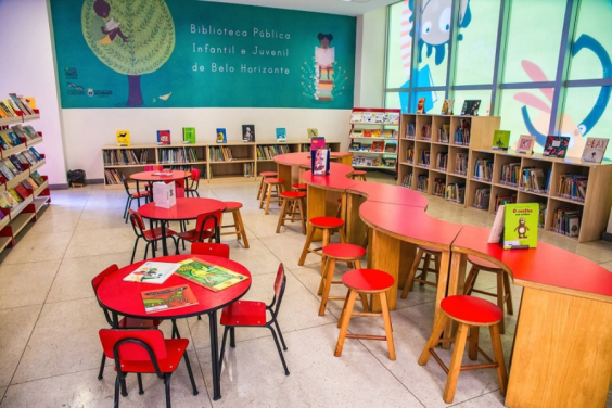 Loca para leitura infantil, com mesa e bancada para diferentes faixas etárias e prateleiras com livros diversos. Ao fundo, parede pintada de verde escuro com ilustração de duas crianças lendo e os seguintes dizeres ao meio: "Biblioteca Pública Infantil e Juvenil de Belo Horizonte".