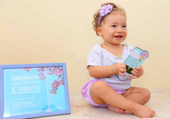 Criança caucasiana usa lado de fita lilás no cabelo, blusa branca e short lilás. ao lado, um quadro com os dizeres: "Amamentar não é só amor. É um direito."