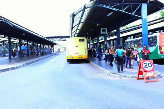 Na Estação Diamante  passageiros, em pé, aguardam o ônibus. Estacionado, um ônibus amarelo aguarda a saída