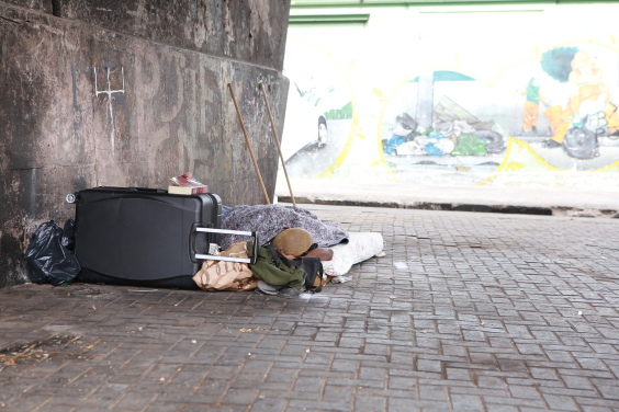Homem com cabelos curtos pintados de amarelo está deitado no chão com um cobertor, ao lado de uma mala e debaixo de um viaduto . Ao fundo, um muro com pinturas urbanas que mostram lixo descartado irregularmente debaixo de um viaduto