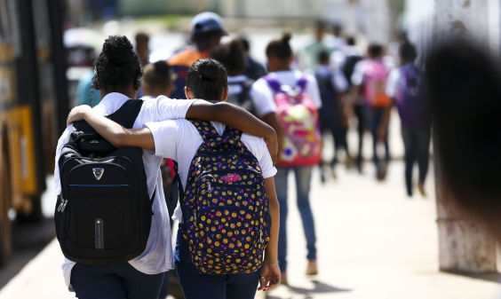 Duas estudantes, de costas, cada uma com uma mochila, abraçadas, caminham, durante o dia. Ao fundo, mais estudantes. 