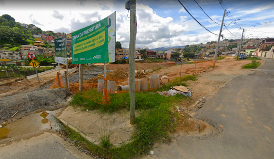 Via de terra com tubulações à direita e casas de comunidade ao fundo. à esquerda, placa com os seguintes dizeres: "Obra da Prefeitura. Tratamento de fundo de vales dos córregos Olaria e Jatobá 3ª etapa". 