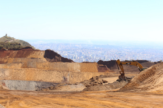 Vista da serra onde a mineração destruiu a paisagem. À direita, um máquina revolve a terra; ao fundo , o céu azul e a cidade de BH