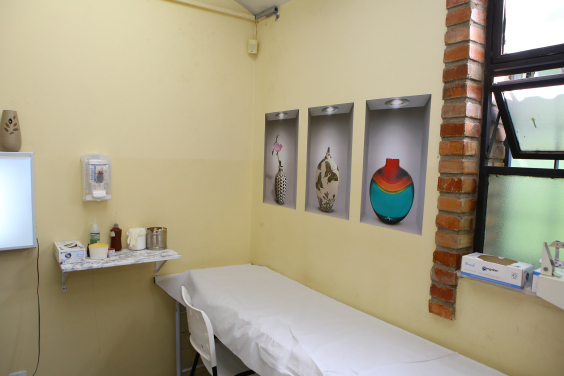 Consultório médico vazio. Uma maca forrada, debaixo da janela aberta, onde estão apoiadas algumas caixas de insumos