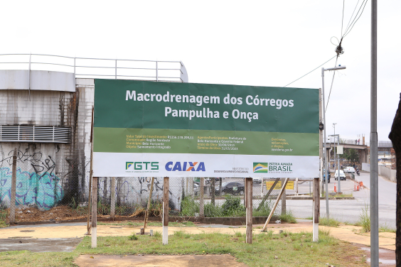 Placa indicativa de obras nos córregos  Pampulha e Onça. Com fundo preto e letras brancas, a placa traz logomarcas do FGTS, da Caixa Econômica Federal, do Ministério  de Desenvolvimento Regional e do Governo Federal 