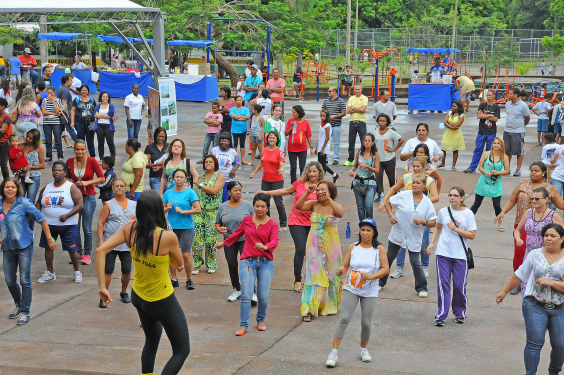 Mais de 40 pessoas praticam atividade física em quadra de escola, orientados por uma mulher, durante o dia.