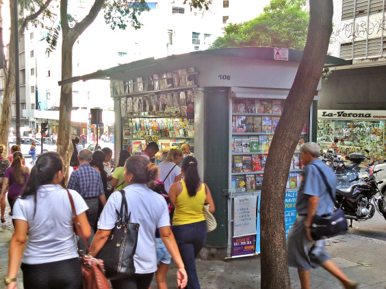 Várias pessoas passam diante de uma banca de revista no centro da cidade