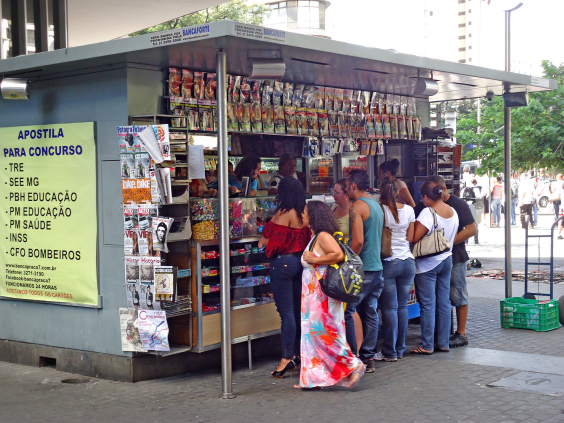 Imagem de banca de revista com clientes fazendo compras. Afixado ao lado da banca, um cartaz informando sobre concursos públicos