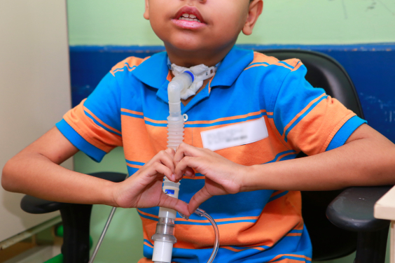 Criança vestida com camisa listrada de azul e laranja, com um aparelho ligado na traqueia, sentado em uma cadeira. Ele faz um gesto de coração com as duas mãos à frente do corpo