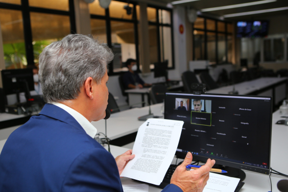 Sentado à mesa, vereador participa de reunião remota, se comunicando com outras pessoas na tela do computador.