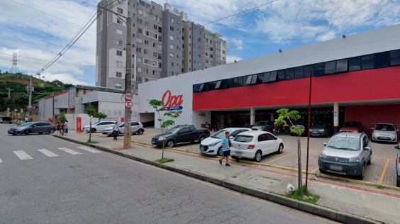 Rua em frente a supermercado, com carros estacionados e faixa para pedestres, durante o dia. 