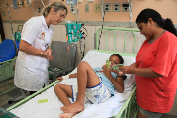 Criança em leito hospitalar acompanhada de mulher e atendida por profissional de saúde.