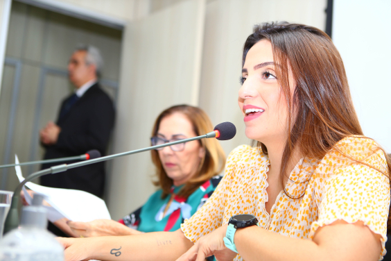 Duas vereadoras sentadas à mesa, em reunião presencial. Ao fundo, de pé, assessor da Câmara.