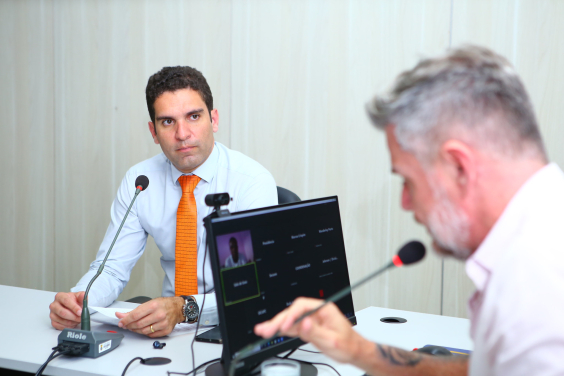 Dois vereadores sentados em reunião presencial e um deles exibido em tela de computador, participando de maneira remota. 