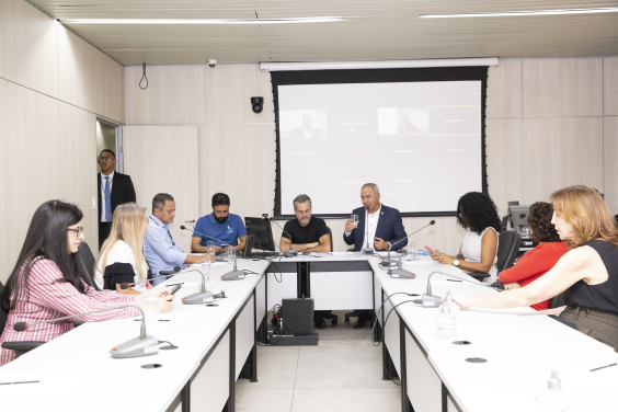 Imagem da mesa em formato de U com participantes da audiência. 