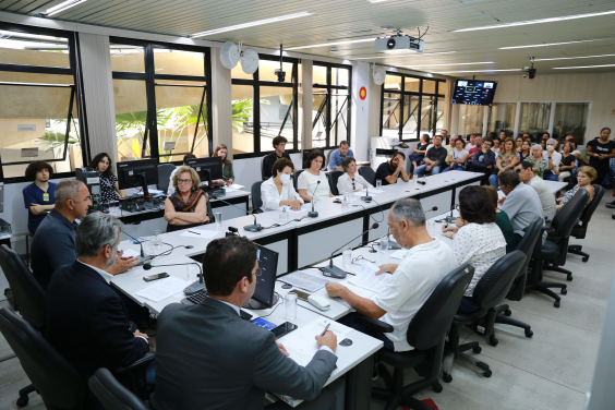 três vereadores à esquerda, dez participantes ao logo da mesa em formato de "u" e mais de vinte cidadãos acompanham audiência pública